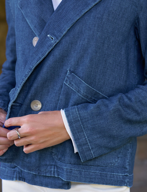 Close-up of a person wearing a blue denim-style double-breasted blazer with white shirt cuffs and a white undershirt visible, plus a silver ring on one finger., view 3