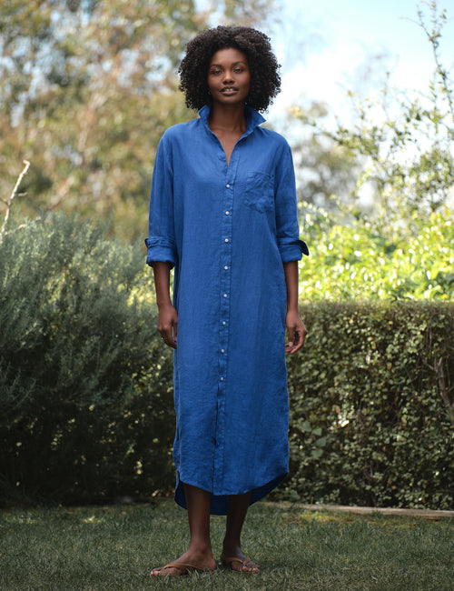 Woman wearing a blue dress standing outdoors with greenery in the background, front view