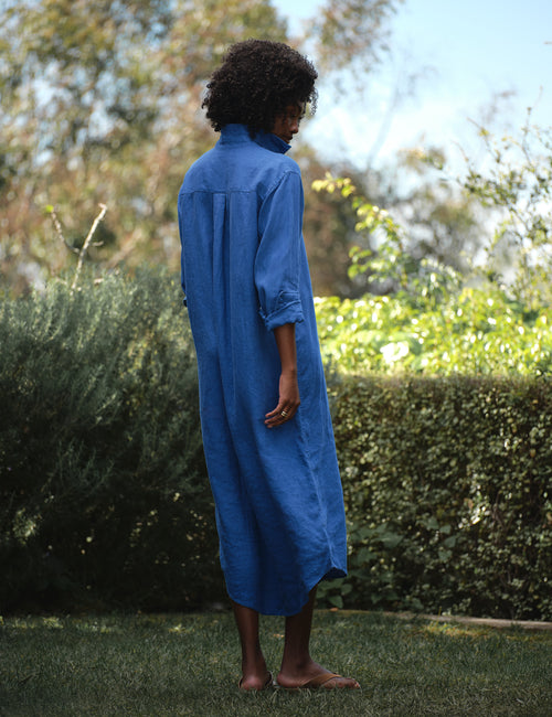 Person wearing a blue dress standing outdoors with greenery in the background, alternate view