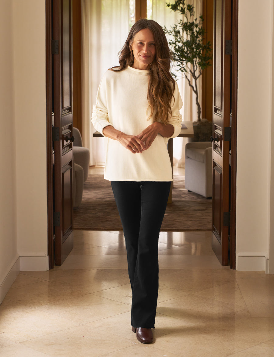 Woman standing in a hallway wearing a white sweater and dark pants.