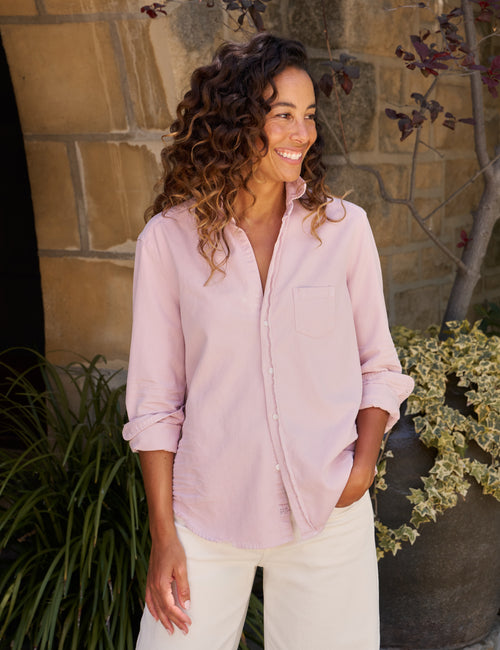 A woman with curly brown hair wearing a light pink button-down shirt and cream pants, standing outdoors in front of a stone wall and green plants., front view