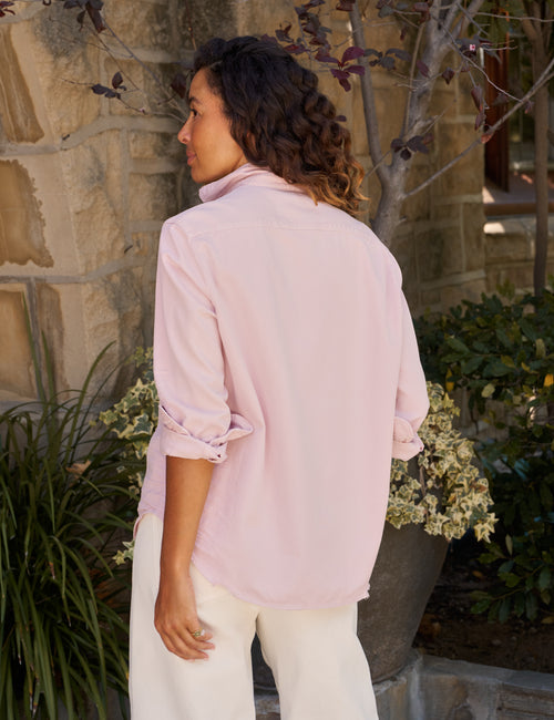 A woman wearing a light pink long-sleeve button-up shirt with rolled-up sleeves and white pants, standing outdoors in front of a stone wall and plants., alternate view