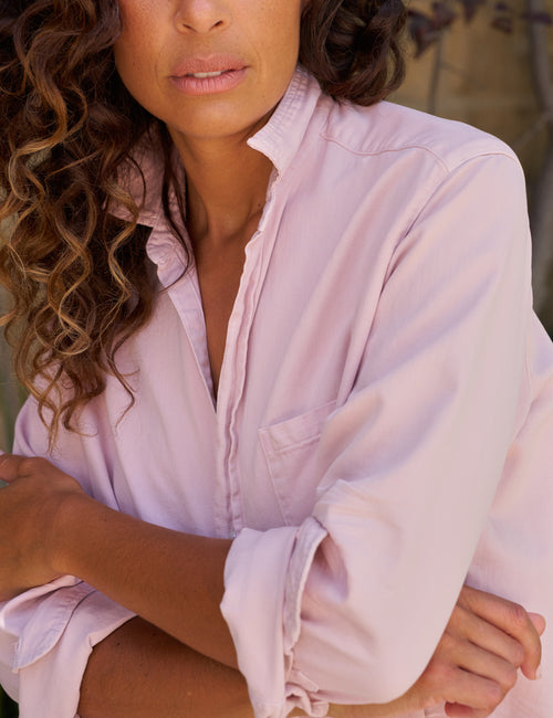 A close-up of a woman wearing a light pink button-up shirt with rolled sleeves, shown from the chest up against a softly blurred outdoor background., view 3