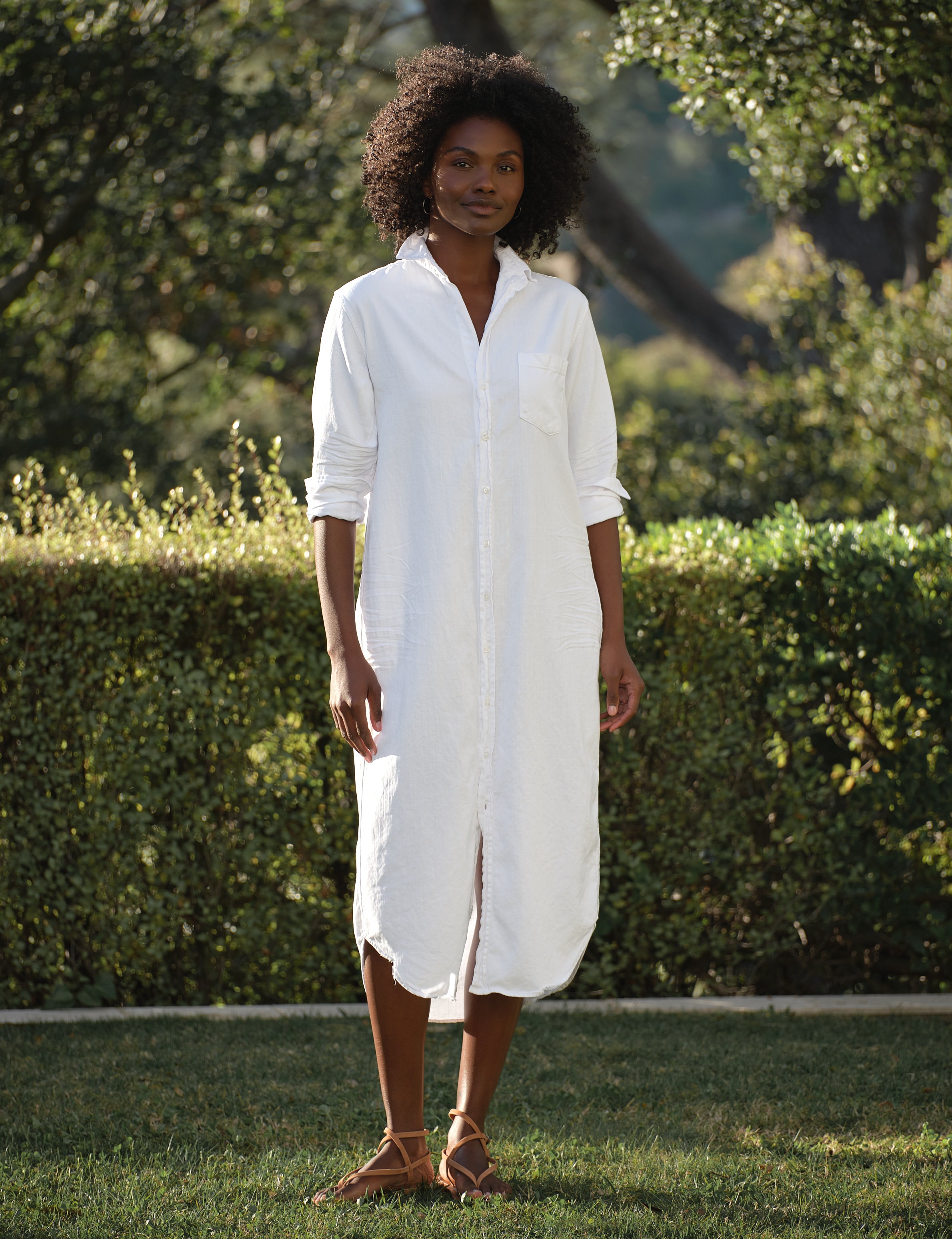 Woman wearing a white dress standing outdoors with greenery in the background, front view
