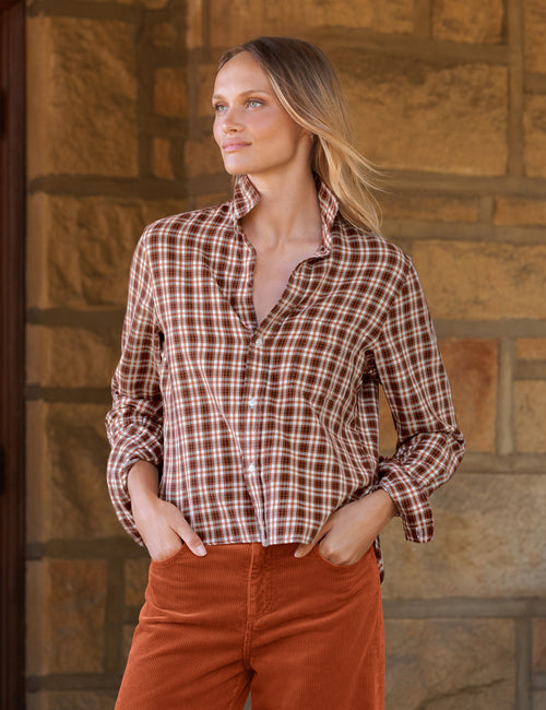 A woman wearing a brown and white plaid button-down shirt and rust-colored pants, standing in front of a stone wall., front view