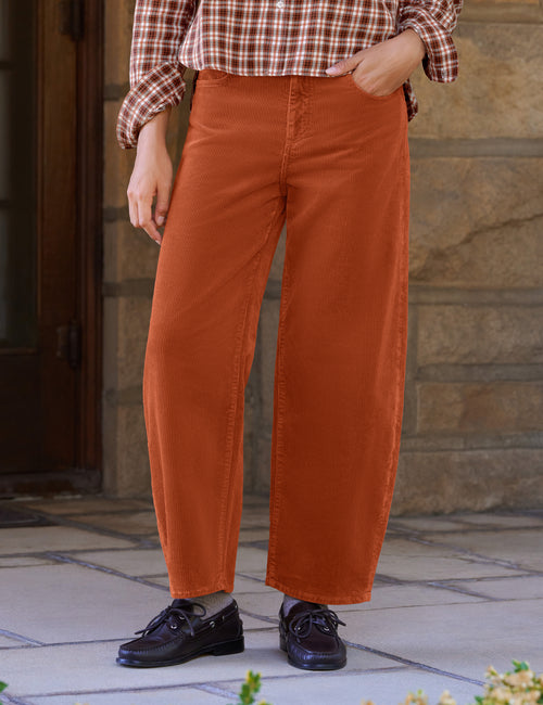 A person wearing rust-orange barrel-leg corduroy pants, a brown and white plaid shirt, and black loafers, standing outdoors on a stone patio., front view