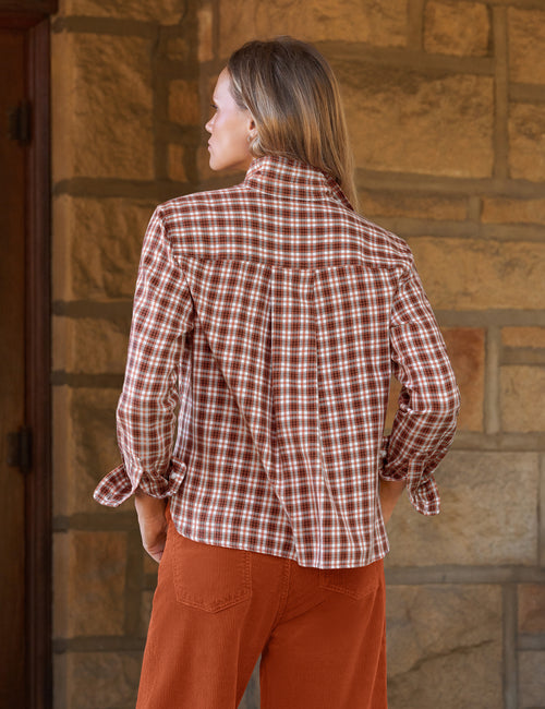 A woman shown from the back wearing a brown and white plaid button-down shirt and rust-colored pants, standing in front of a stone wall., alternate view