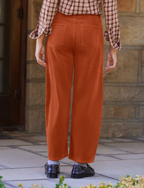 A person shown from the back wearing rust-orange barrel-leg pants, a brown and white plaid shirt, gray socks, and black loafers, standing outdoors in front of a stone wall., view 3