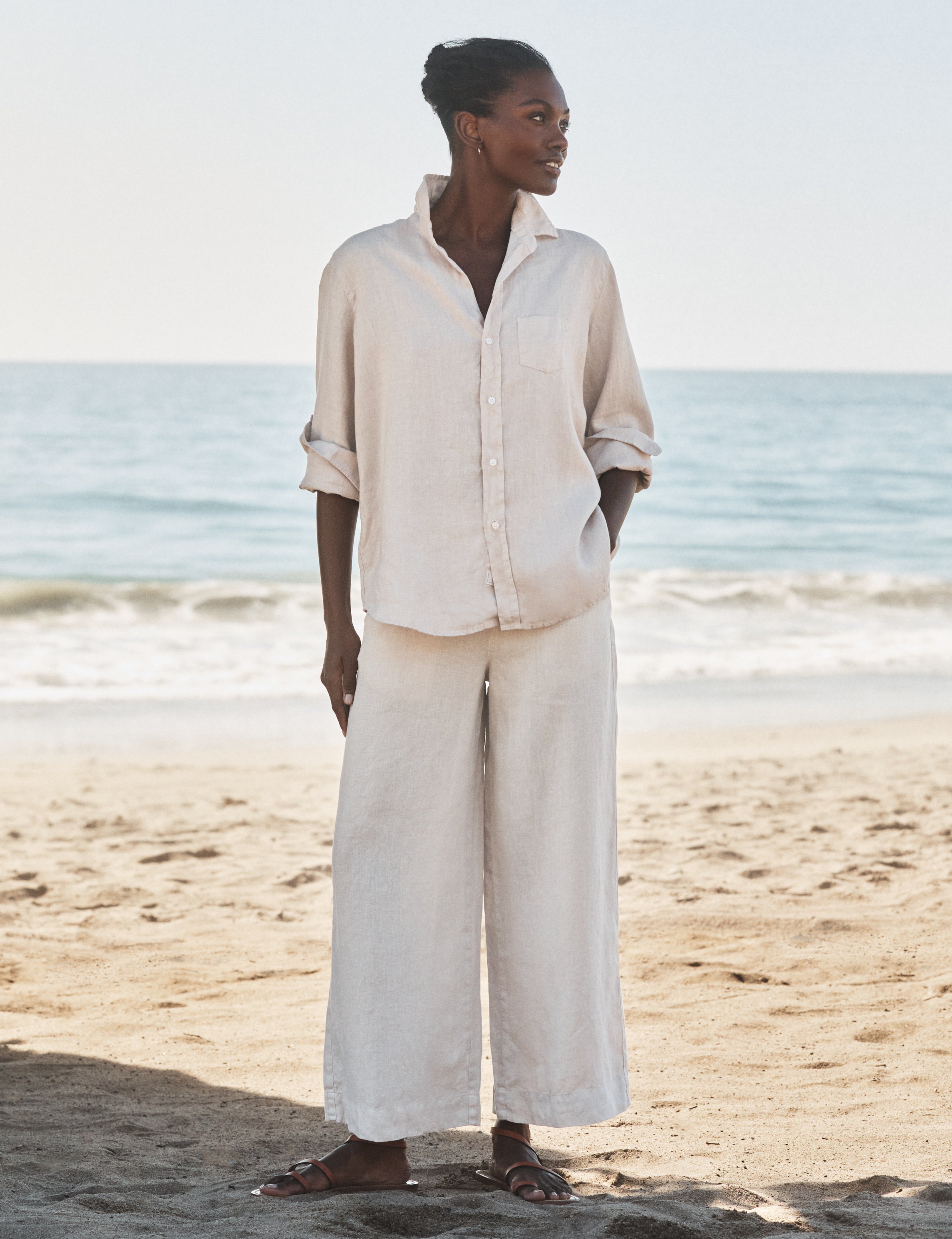 Woman in beige outfit standing on a beach with ocean in the background, front view