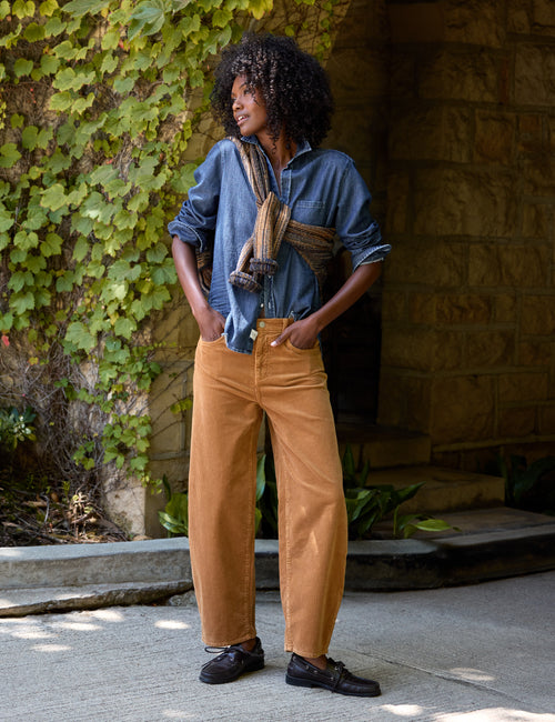 A person standing outdoors in front of a stone wall and ivy-covered wall, wearing a blue button-up shirt, tan corduroy pants, and black shoes, with a brown-and-beige sweater wrapped around the shoulders., alternate view