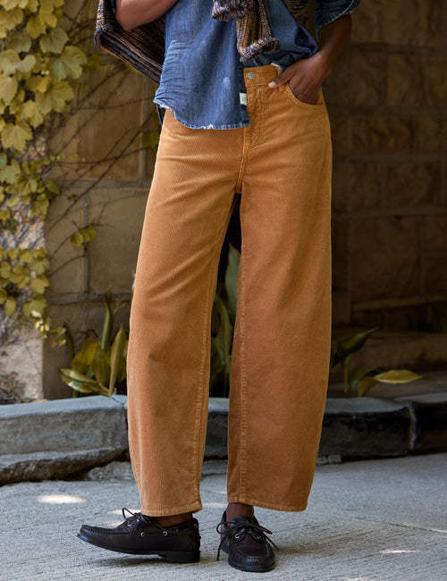 A person wearing wide-leg tan corduroy pants, a blue denim shirt, and black lace-up shoes, standing outdoors against a stone wall with greenery., front view