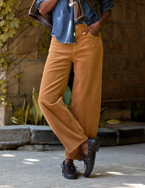 A person wearing barrel-leg tan corduroy pants, a blue denim shirt, and black shoes, standing outdoors against a stone wall and plants., view 4
