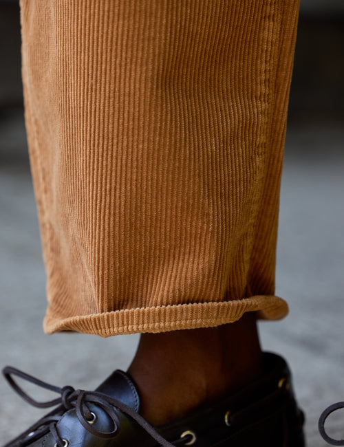 Close-up of a person's ankle wearing cuffed tan corduroy pants and a black leather lace-up shoe, with a blurred gray concrete background., view 3