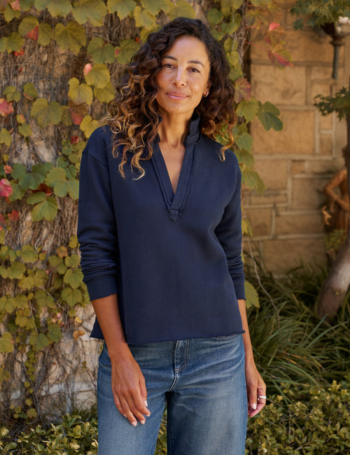A woman with curly brown hair wearing a navy blue long-sleeve  top and blue jeans, standing outdoors in front of a stone wall and ivy-covered tree., front view