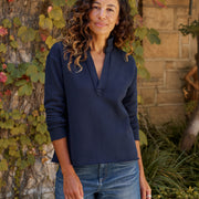 A woman with curly brown hair wearing a navy blue long-sleeve  top and blue jeans, standing outdoors in front of a stone wall and ivy-covered tree.
