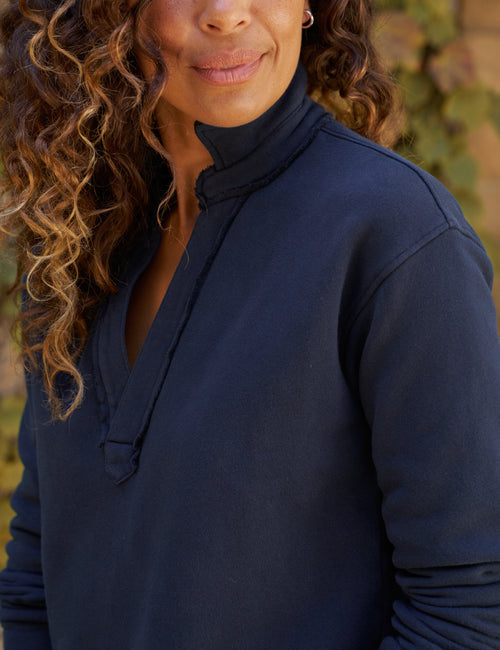 A close-up of a woman wearing a navy blue pullover sweatshirt, showing the fabric texture and collar detail against a soft, blurred outdoor background., view 3