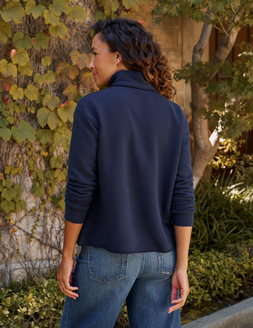A woman wearing a navy blue long-sleeve top, shown from the back in an outdoor setting with ivy-covered walls and trees., alternate view