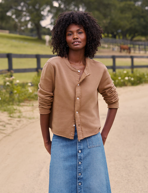 A woman with curly black hair stands outdoors on a dirt path wearing a cropped tan button-front cardigan and a blue denim skirt. A fence, grass, and trees are blurred in the background., front view