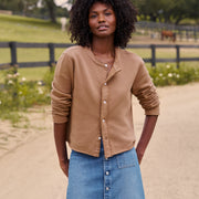 A woman with curly black hair stands outdoors on a dirt path wearing a cropped tan button-front cardigan and a blue denim skirt. A fence, grass, and trees are blurred in the background.
