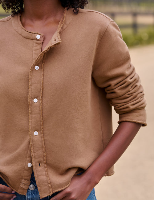 A close-up of a person wearing a tan long-sleeve button-front top with raw, frayed edges and white buttons, photographed outdoors against a blurred light brown background., view 3