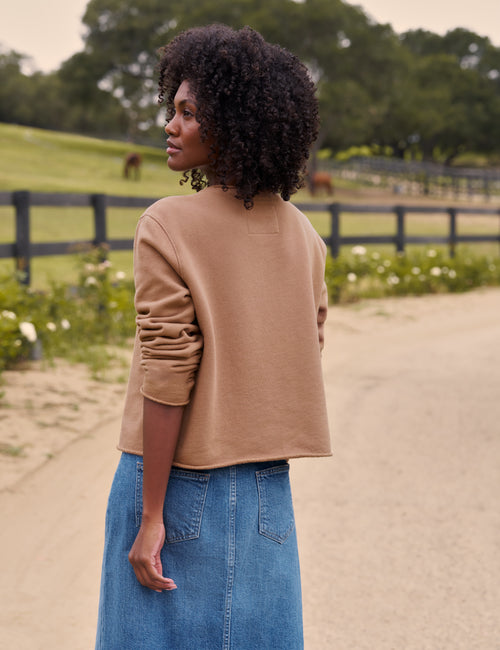 A woman with curly black hair is shown from the back wearing a cropped tan long-sleeve knit top and a blue denim skirt, standing on a dirt path in a rural outdoor setting with a fence and green fields in the background., alternate view