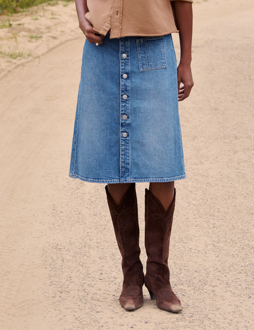 A woman stands on a dirt path in a rural outdoor setting, wearing a tan cropped button-front cardigan, a blue denim midi skirt, and brown knee-high boots., front view