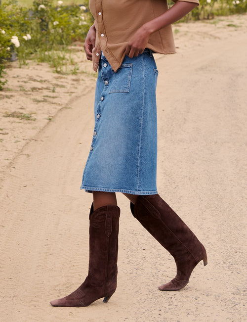 A woman walking on a dirt path outdoors, wearing a tan button-front cardigan, a light blue denim midi skirt, and dark brown heeled boots. A fence, green field, and trees are visible in the background., alternate view