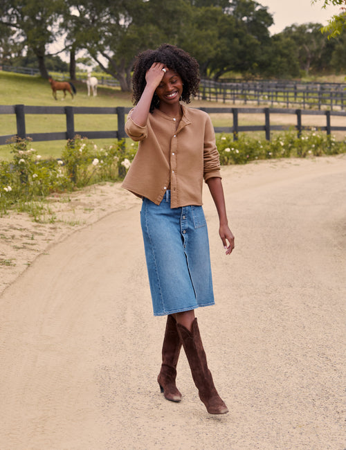 A woman stands on a dirt path outdoors wearing a tan cropped button-front cardigan, a blue denim midi skirt, and brown knee-high boots. A fence, green field, and trees are visible in the background., view 3