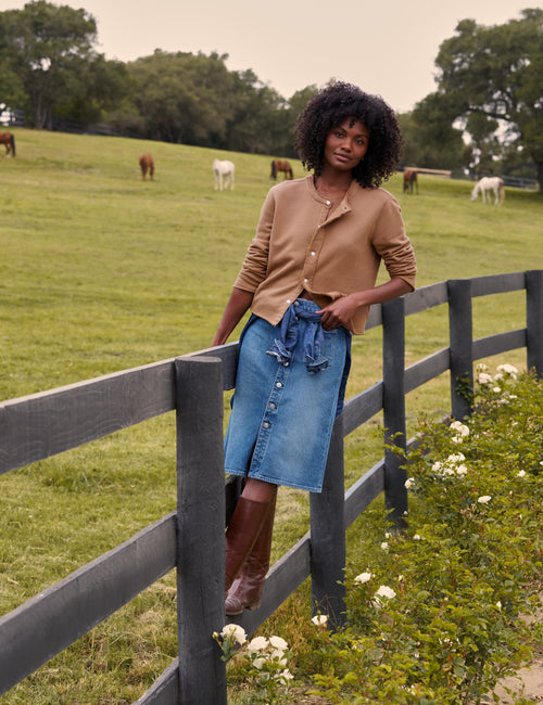A woman stands outdoors by a wooden fence in a grassy field with cows in the background. She is wearing a tan button-front cardigan, a blue denim skirt, and brown knee-high boots., view 5