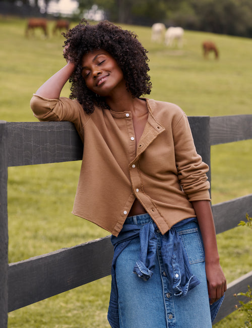 A woman with curly dark hair leans against a dark wooden fence in a grassy field, wearing a cropped tan button-front cardigan over a brown top and a blue denim skirt., view 4