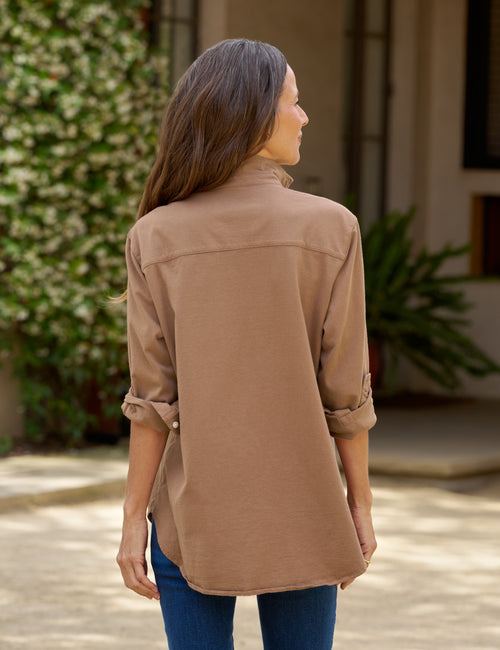 A woman viewed from the back wearing a tan long-sleeve button-up shirt with rolled-up sleeves and blue jeans, standing outdoors in front of a building with greenery., alternate view
