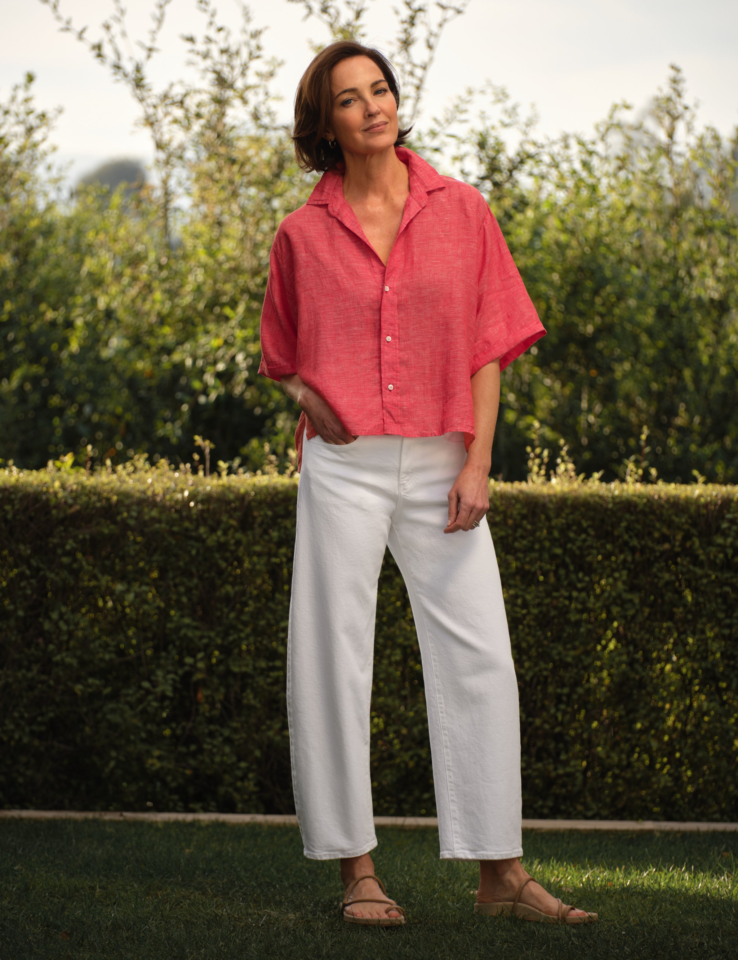 Woman wearing a red shirt and white pants standing outdoors with greenery in the background, view 4
