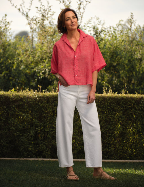 Woman wearing a red shirt and white pants standing outdoors with greenery in the background, view 4