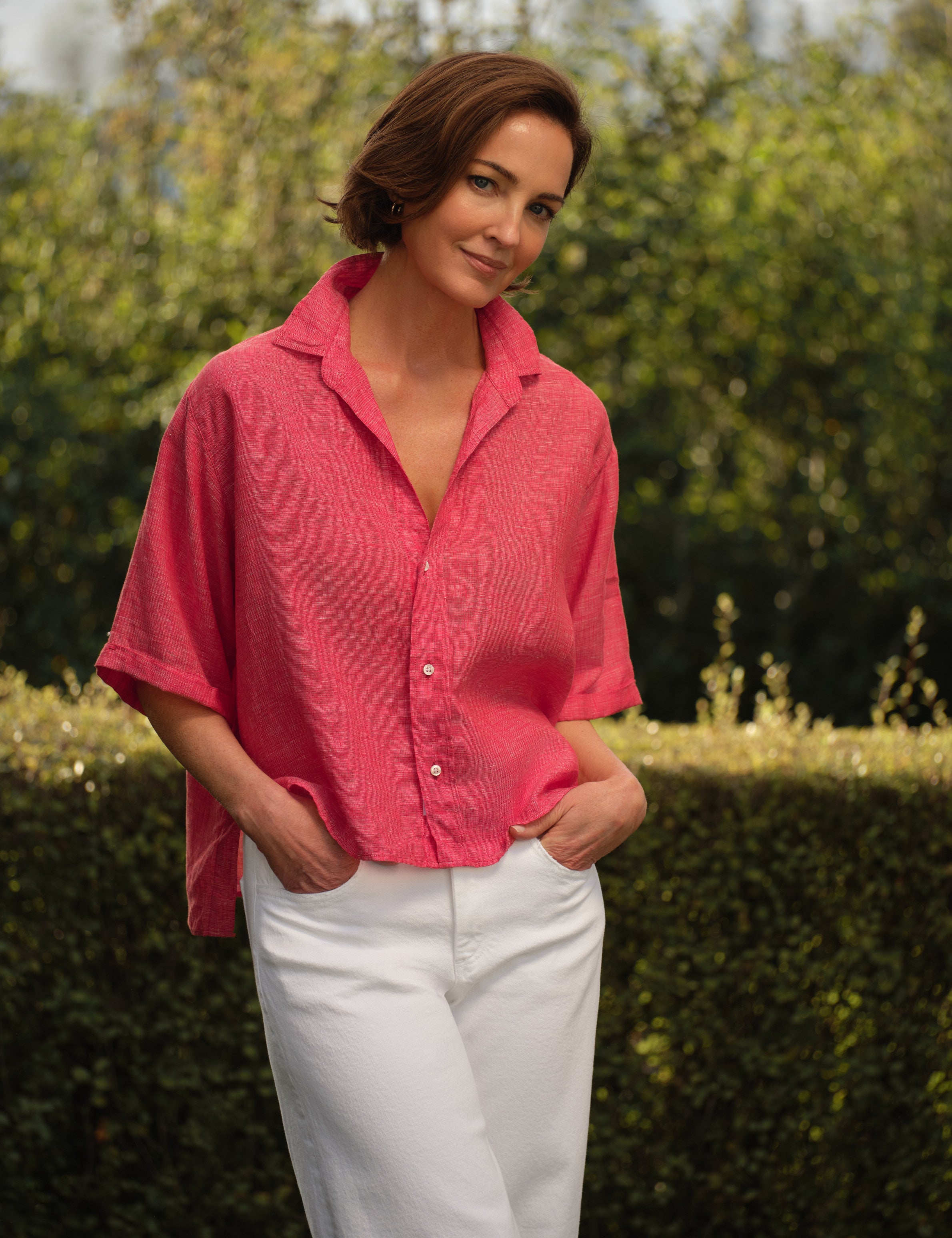 Woman wearing a red shirt and white pants standing outdoors with greenery in the background, front view
