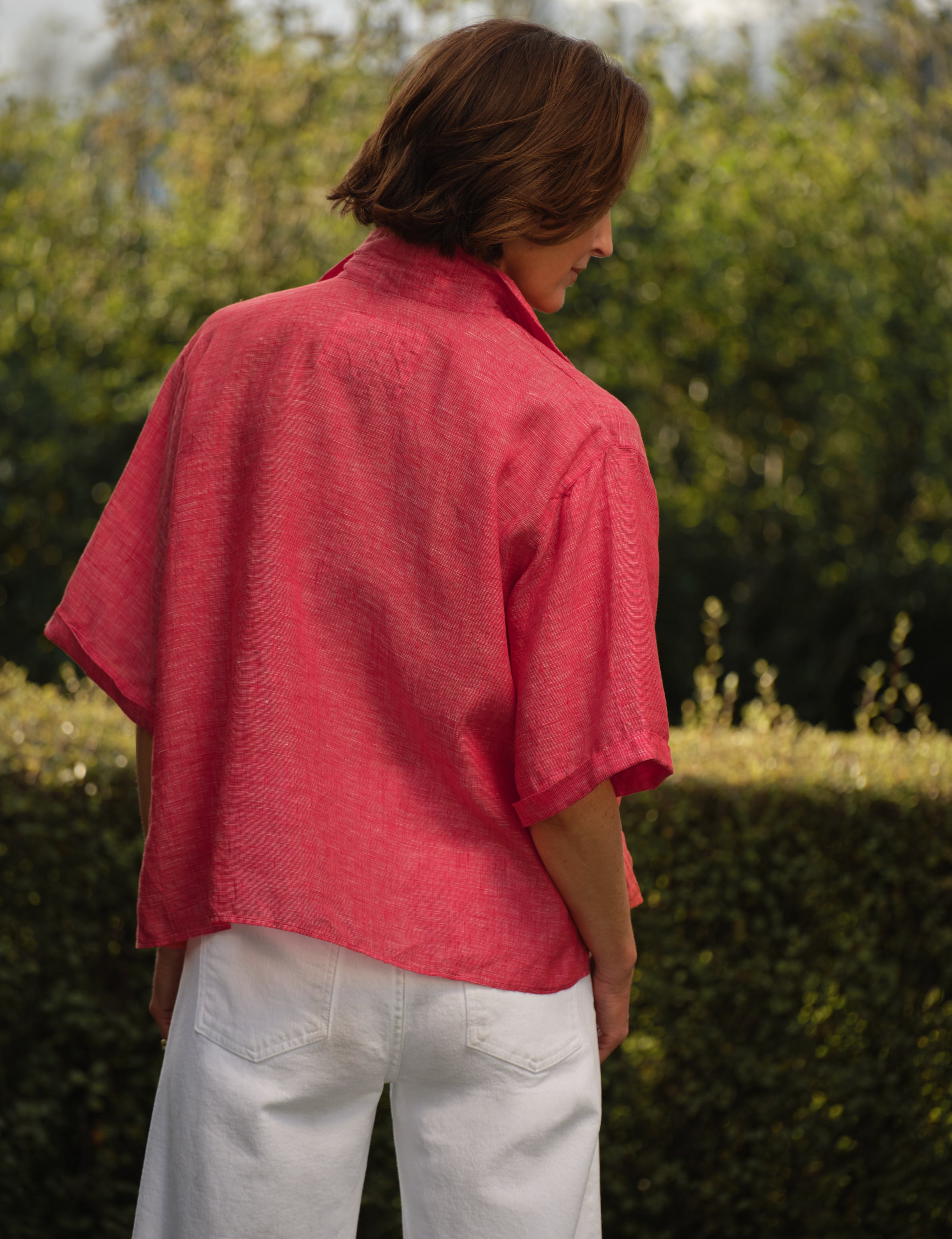 Person wearing a red shirt with a brand logo, standing outdoors with greenery in the background, alternate view