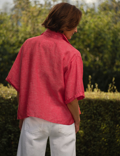Person wearing a red shirt with a brand logo, standing outdoors with greenery in the background, alternate view