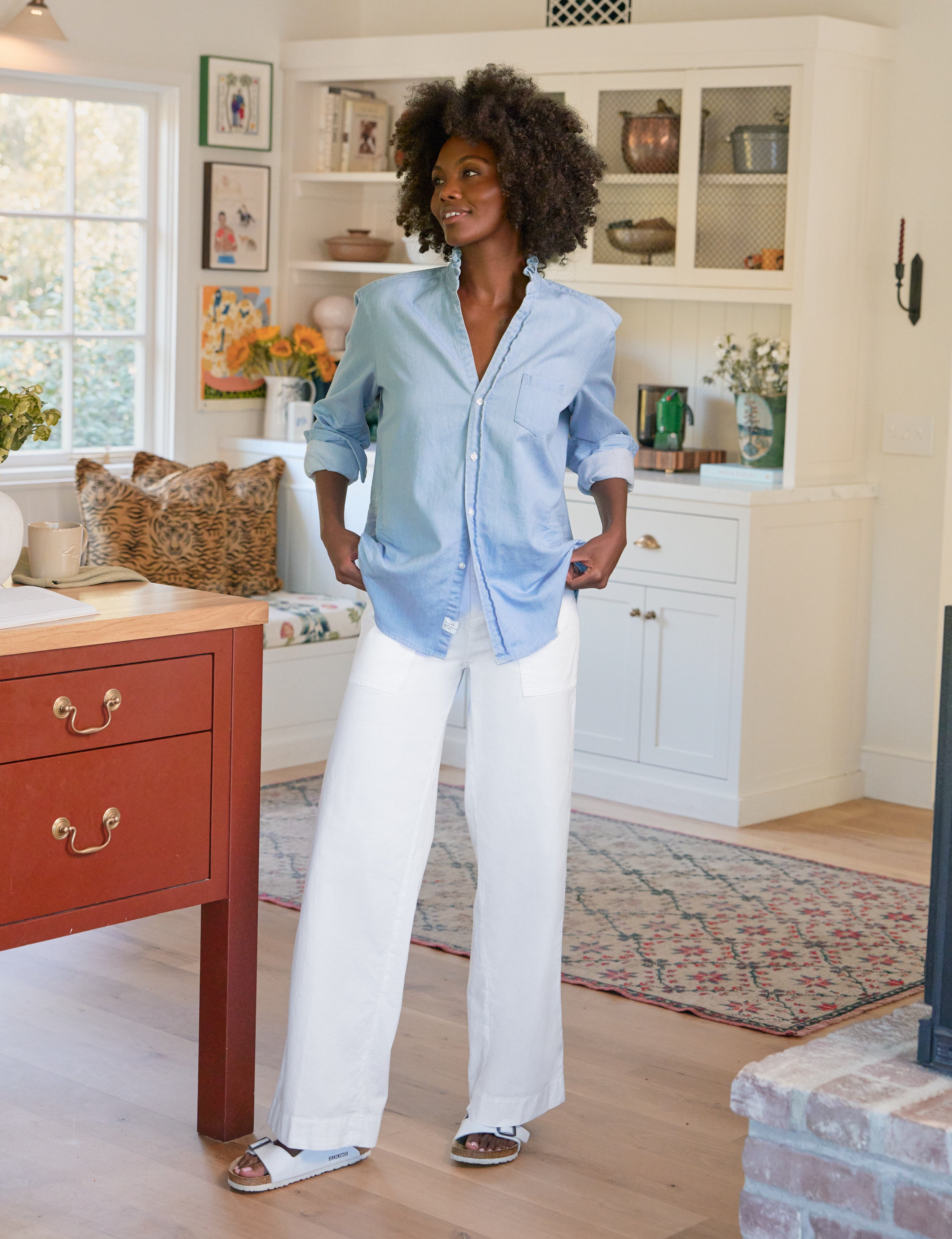 Woman standing in a kitchen wearing a light blue shirt and white pants.