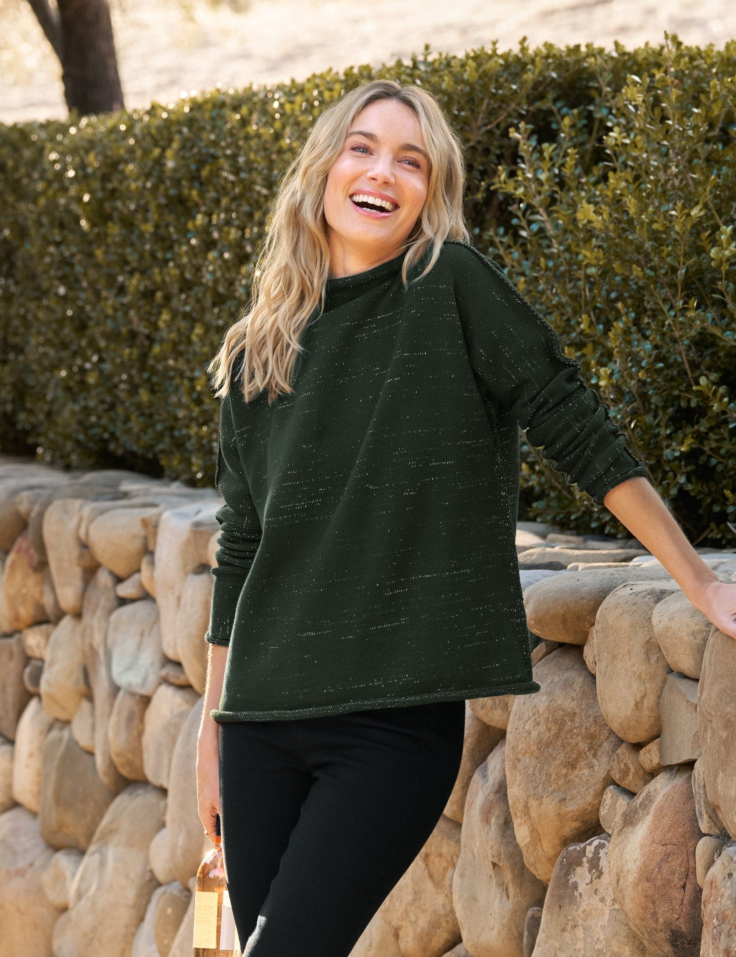 Woman in a green sweater and black pants standing next to a stone wall outdoors.