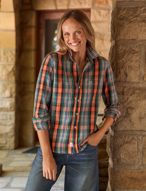 A woman smiling and leaning against a stone pillar, wearing a long-sleeve button-up plaid shirt in orange, green, and blue tones, paired with blue jeans., front view