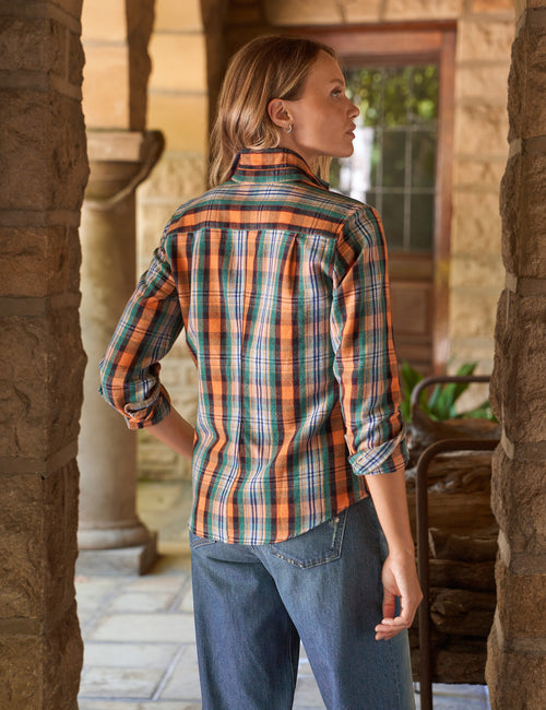 A woman wearing a long-sleeve plaid button-up shirt in orange, green, and blue, paired with blue jeans, standing in a stone corridor., alternate view