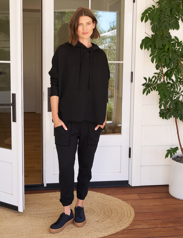 A woman standing in front of a door, wearing a black hoodie and black joggers from the Venice Travel Set, with her hands in the pockets of the joggers.