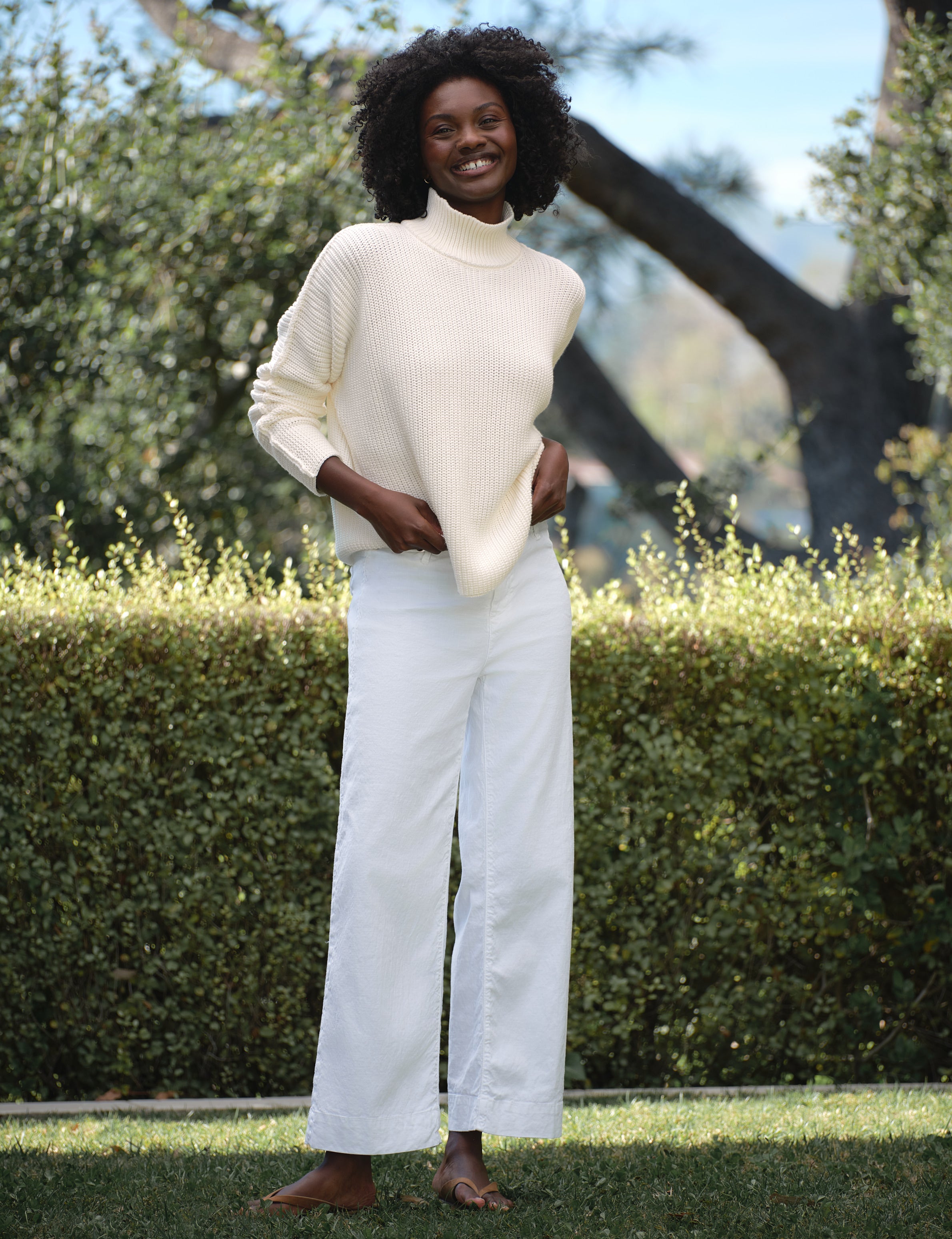Person wearing a cream turtleneck sweater and white pants standing outdoors with greenery in the background, view 5