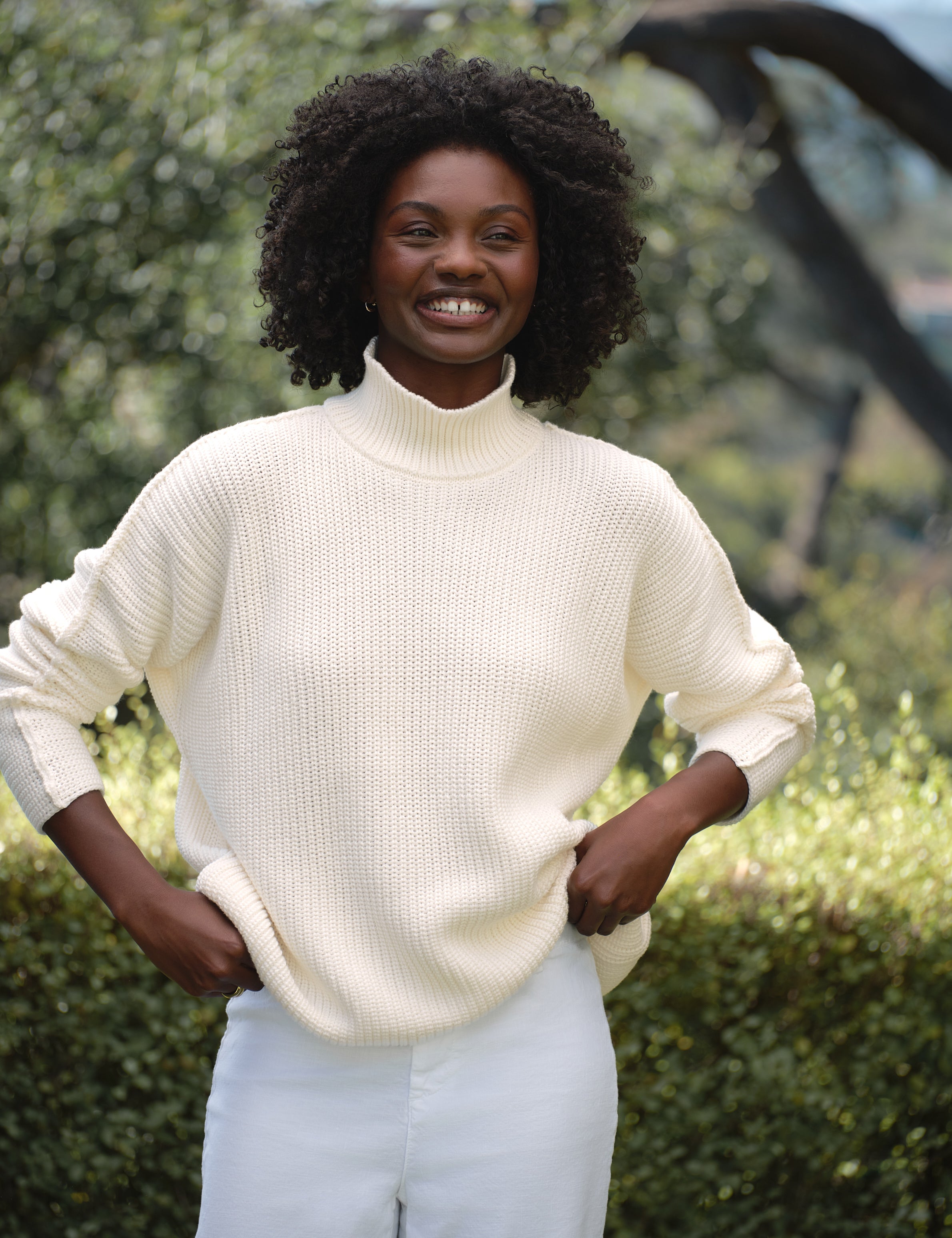 Woman wearing a white sweater and white pants standing outdoors with greenery in the background, front view
