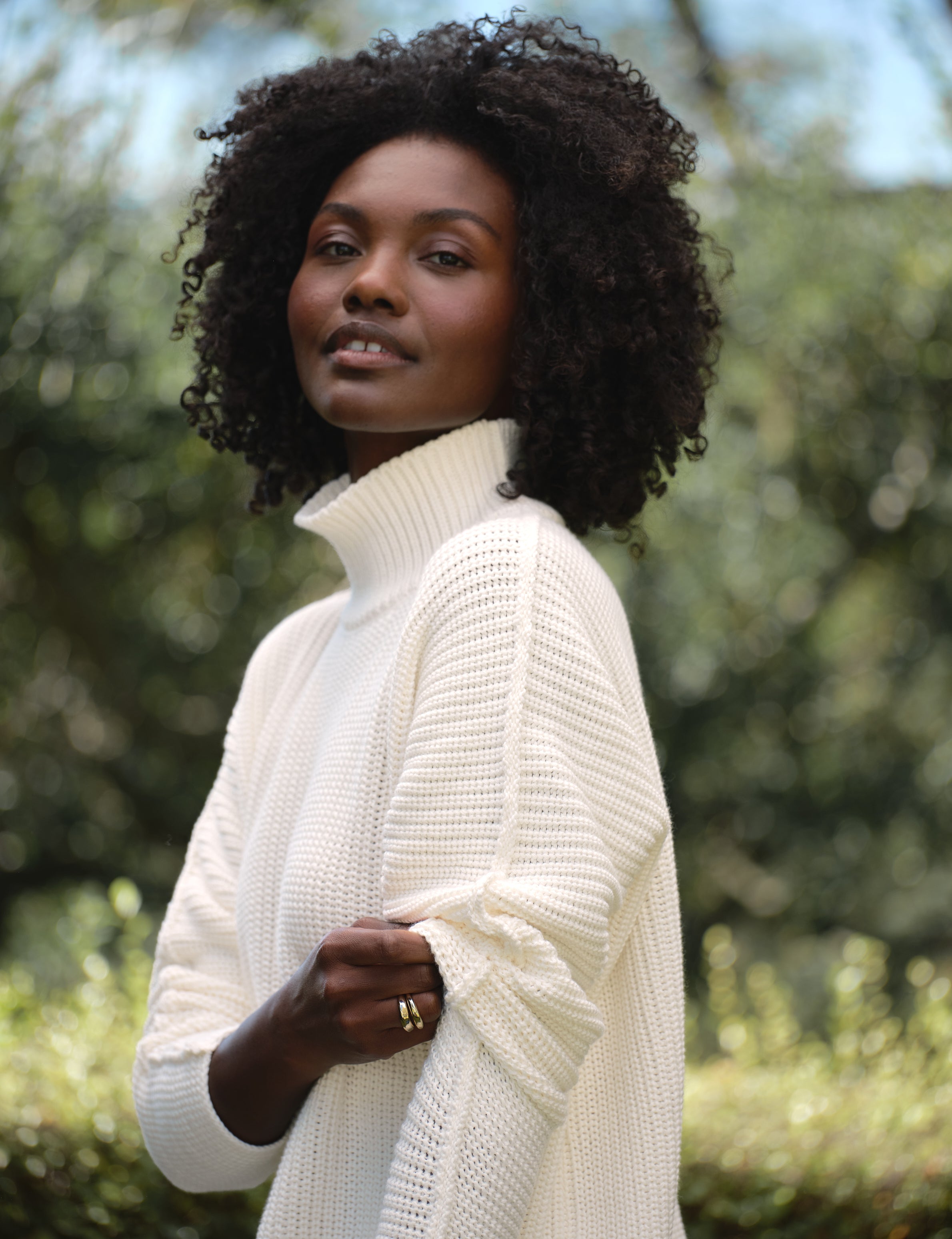 Woman wearing a white turtleneck sweater outdoors with greenery in the background, view 4