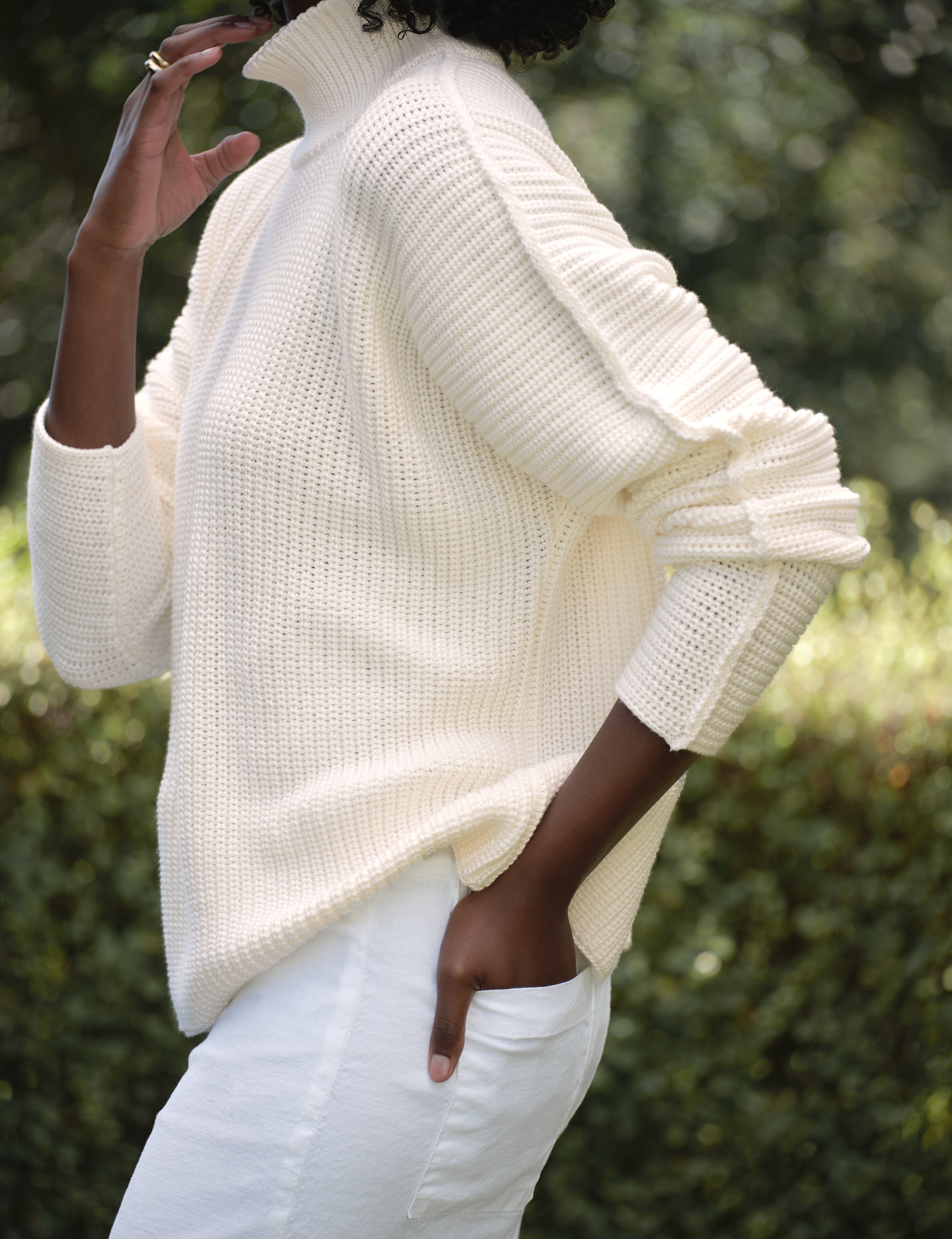 Person wearing a white sweater and white pants with a blurred green background, view 3