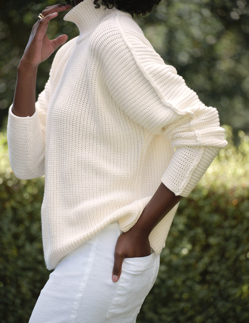 Person wearing a white sweater and white pants with a blurred green background, view 3
