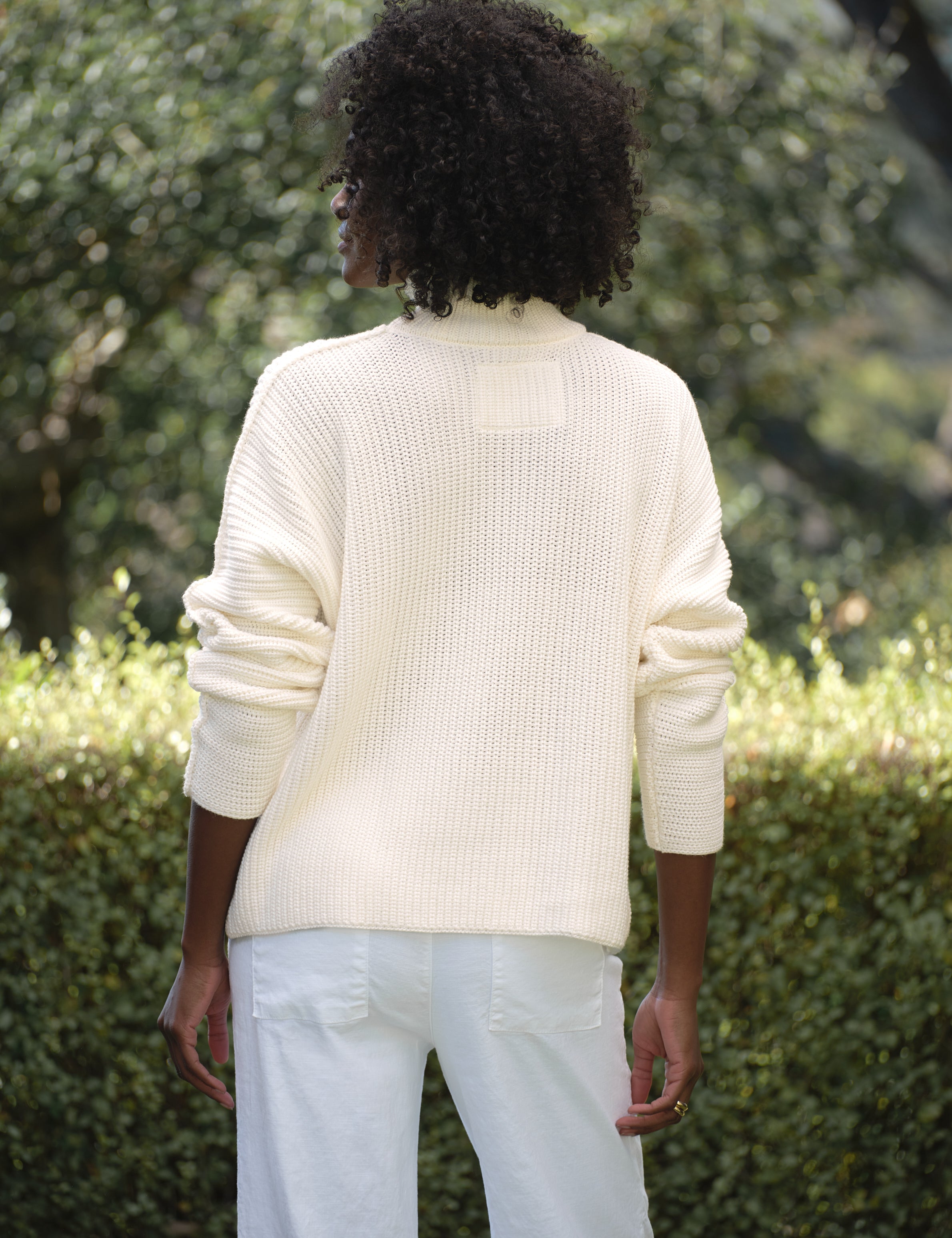 Person wearing a cream sweater and white pants standing outdoors with greenery in the background, alternate view