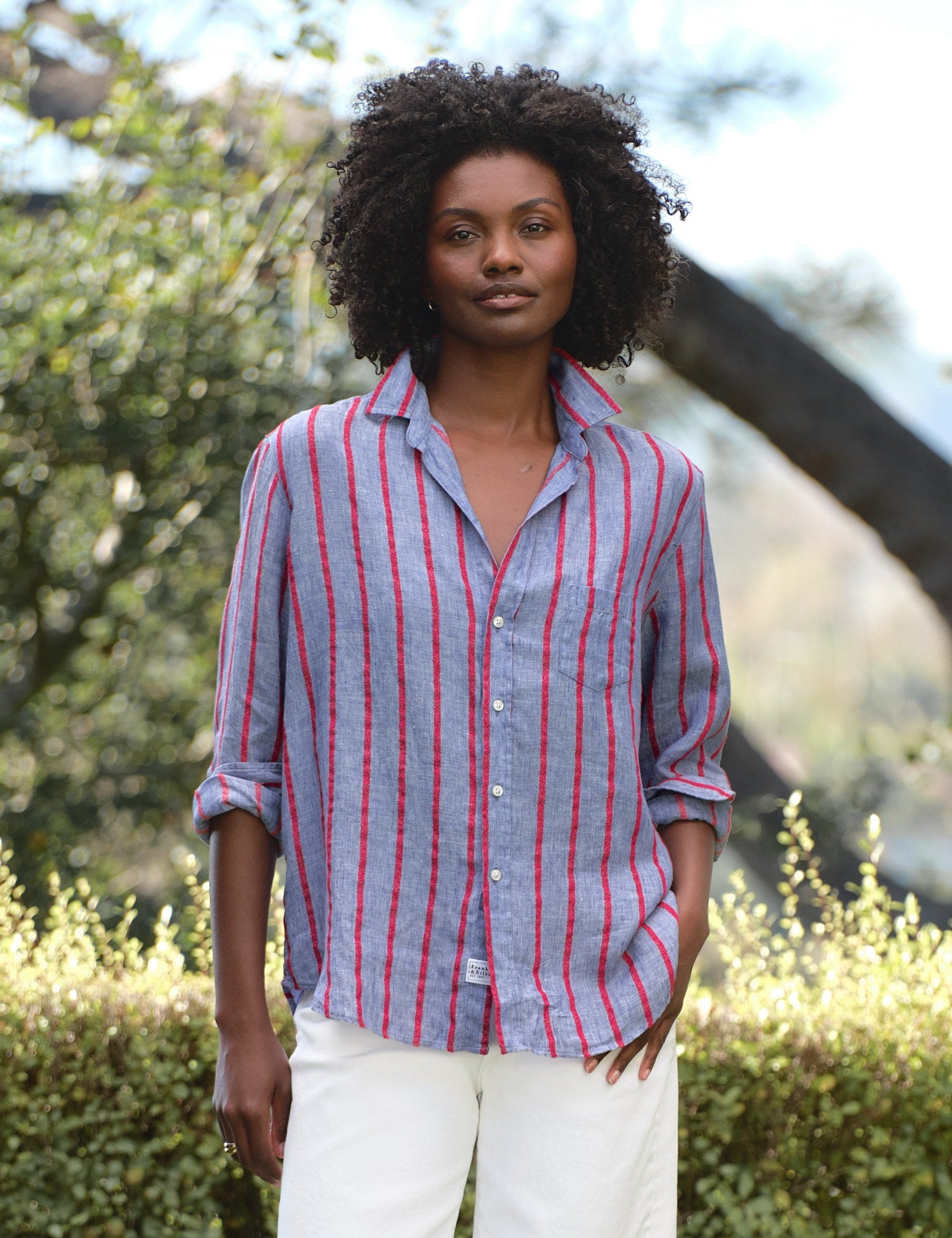 Woman wearing a striped shirt and white pants standing outdoors with greenery in the background, view 4