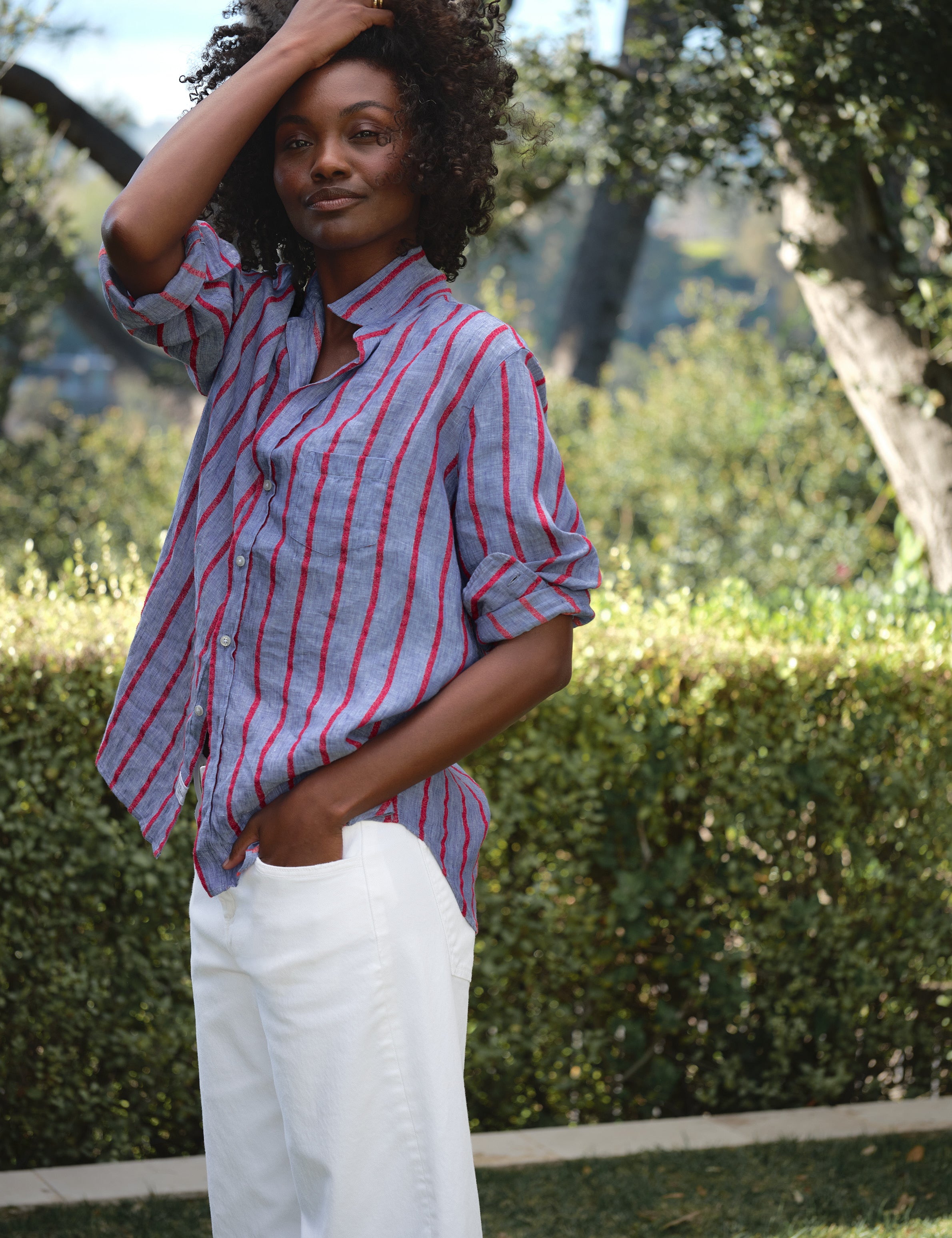 Woman wearing a red and blue striped shirt and white pants standing outdoors with greenery in the background, view 5