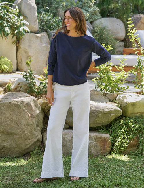 Woman wearing a navy top and white pants standing in a garden with rocks and plants., view 4